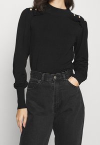 Czarny sweter z dzianiny o wysokim dekolcie, z falbanami i perłowymi akcentami na ramionach, w połączeniu z czarnymi dżinsami o wysokim stanie.