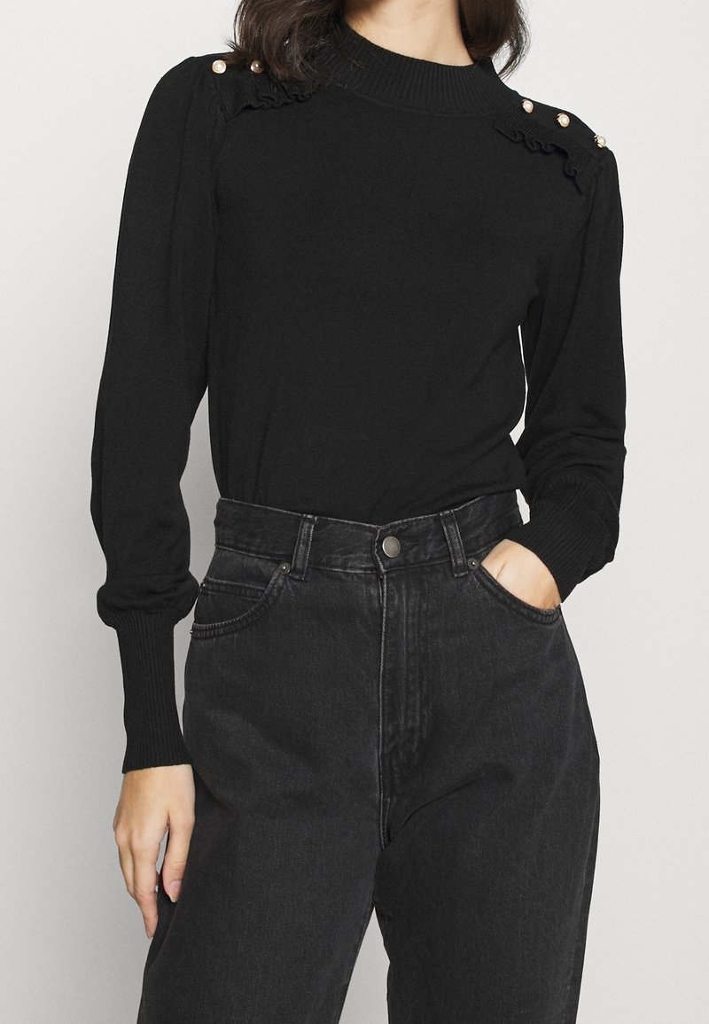 Czarny sweter z dzianiny o wysokim dekolcie, z falbanami i perłowymi akcentami na ramionach, w połączeniu z czarnymi dżinsami o wysokim stanie.