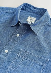 Chemise en jean bleu à boutons avec col pointu, boutons blancs, poche avant et étiquette de marque visible à l'intérieur du col.