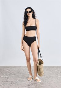 Σετ μαύρου μπικίνι με τοπ μπαντό και ψηλόμεσα κάτω μέρη. Το μοντέλο φοράει λευκά σανδάλια τύπου σλάιντ και κρατά μια πλεκτή ψάθινη τσάντα. Λείο υλικό.