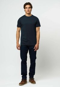 Vyras dėvi tamsiai mėlynį marškinėlį, tamsius džinsus ir rudus odinius batus, stovintis priešais paprastą baltą foną.