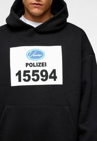 Hoodie noir avec une poche frontale ; présente un patch rectangulaire blanc affichant "POLIZEI" et le numéro "15594" en lettres noires en gras.
