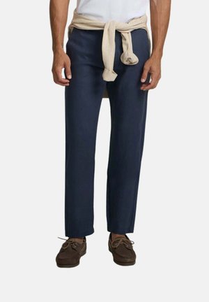 Hombre con pantalones azul marino, camisa blanca, suéter beige atado alrededor de la cintura y zapatos marrones con cordones sobre un fondo liso.