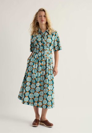 Šviesiaplaukė moteris dėvi žalią midi suknelę su gėlių raštu, trumpomis rankovėmis ir apykakle, ruda mediniais klumpėmis, stovinti priešais vienodą foną.