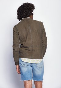 Olivgrüne Lederjacke mit strukturiertem Finish, Reißverschlussdetails, gerippten Bündchen und tailliertem Design. Getragen über einem blumigen Hemd und Jeansshorts.