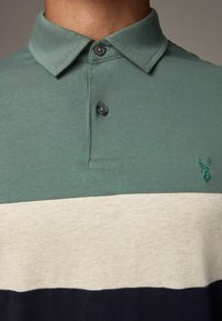 Pehmeast kangast polo-särk, millel on roheline ülemine osa, kreemikas keskriba ja tumesinine alumine osa. Märkimisväärsed mustad nööbid ja tikitud emblem.