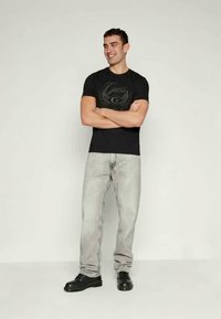 Must must särk logoga, heledad hallid teksad ja mustad jalatsid. Mudel seisab käed risti lihtsa tausta ees.