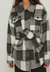 Black and white checked jacket made of wool blend, featuring flap pockets, a belt, and black buttons. The texture appears soft and warm.