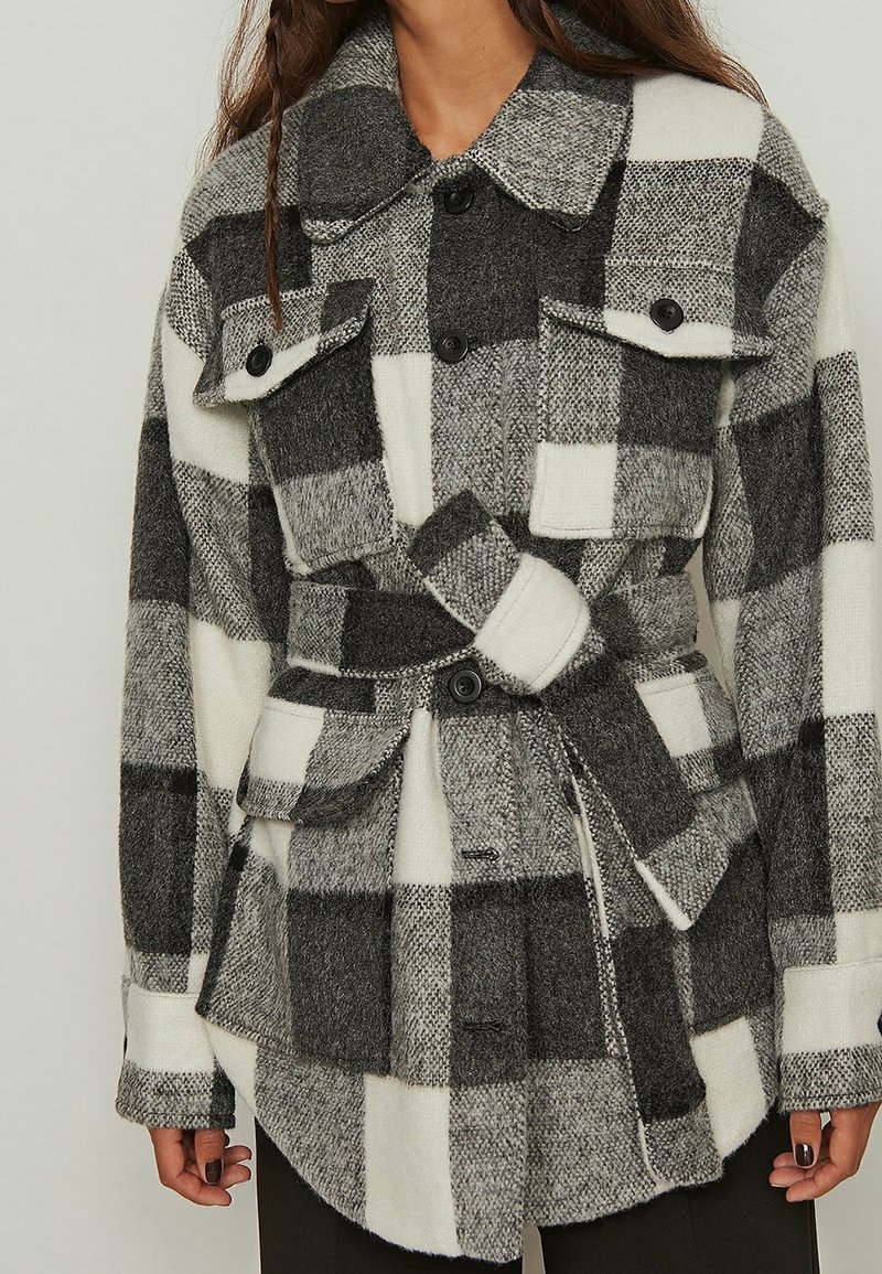 Black and white checked jacket made of wool blend, featuring flap pockets, a belt, and black buttons. The texture appears soft and warm.