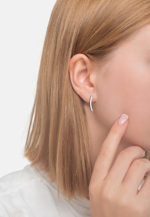 Böjd silverörhänge med en slät yta, bärs på vänster öra. Ett finger rör försiktigt vid kinden. Ljust hår.