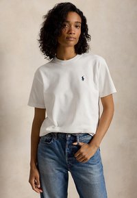Vrouw met krullend haar die een wit T-shirt met korte mouwen draagt met een klein logo op de borst en een blauwe spijkerbroek, poserend met één hand in de zak.