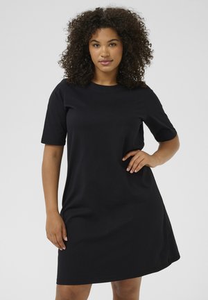 Femme aux cheveux bouclés portant une robe noire unie à manches courtes, debout avec une main sur la hanche sur un fond clair.