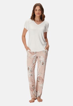 Femme debout pieds nus, portant un haut blanc à manches courtes et un pantalon de pyjama rose clair à motif floral, les mains dans les poches.