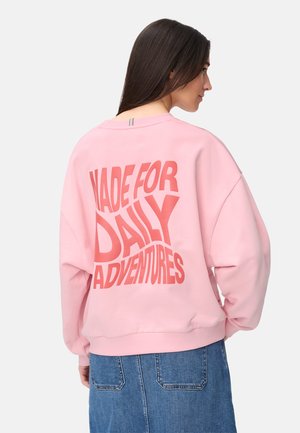 Frau mit langen dunklen Haaren trägt ein pinkes Sweatshirt mit dem Text "MADE FOR DAILY ADVENTURES" auf dem Rücken und einen blauen Jeansrock.