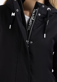 Sort vandtæt jakke med frontlynlås, sølvhardware, justerbar hætte og snørrelukning. Har mærketape langs lynlåsen.