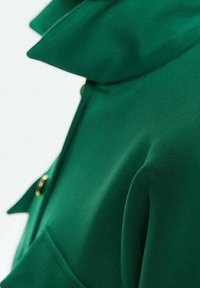 Zielony materiałowy ubiór z wysokim kołnierzem, o gładkiej teksturze i złotym guzikiem w okolicach szyi. Zbliżenie podkreśla detale projektu.