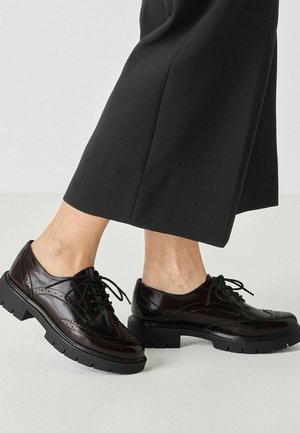 Personne portant un pantalon noir court et des chaussures brogue marron foncé brillantes à lacets avec des semelles noires épaisses marchant sur un sol clair.
