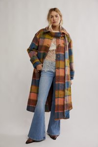 Mulher vestindo um casaco xadrez longo multicolorido, blusa branca de renda, jeans azuis com boca flare e sapatos marrons pontiagudos, em pé contra um fundo simples.