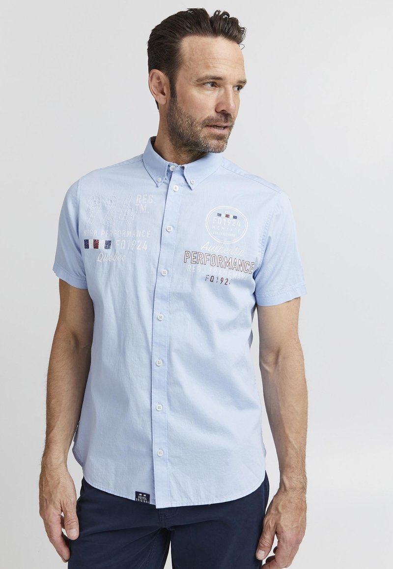Camisa de botones de manga corta azul clara con texto y logotipos bordados, fabricada en un tejido suave y con un diseño de cuello clásico.