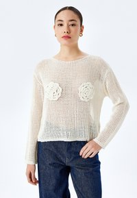 Strickpullover in Creme mit offenem Gewebe und langen Ärmeln, mit zwei strukturierten Blumenapplikationen auf der Brust. Kombiniert mit blauen Jeans.