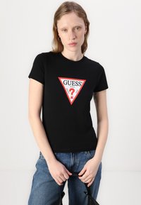 T-shirt en coton noir avec un graphique triangulaire rouge et blanc portant le texte "GUESS ?". Les manches courtes et le design ajusté soulignent la silhouette.
