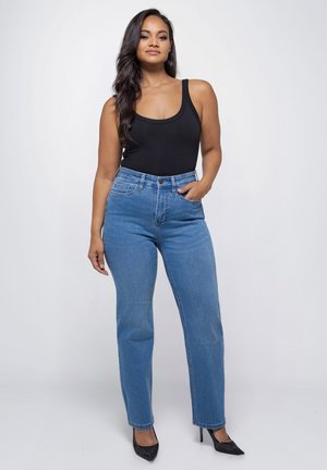 Vrouw staand in een blauwe jeans met hoge taille, zwart mouwloos topje, zwarte hoge hakken, met één hand in de zak tegen een effen achtergrond.