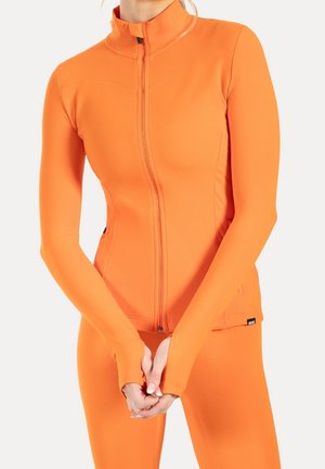 Nahaufnahme einer Person, die eine eng anliegende, leuchtend orangefarbene Reißverschlussjacke mit Daumenlöchern und passende Leggings vor einem einfarbigen Hintergrund trägt.