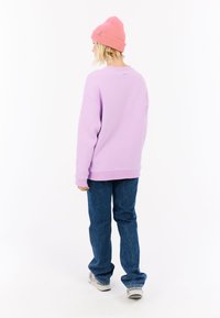 Persona con un gorro rosa, sudadera lavanda, vaqueros azules y zapatillas blancas, de pie de espaldas sobre un fondo blanco.