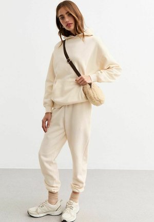 Haut à capuche couleur crème avec une poche kangourou, associé à un pantalon de survêtement assorti. Porté avec un petit sac à texture de fourrure et des baskets.
