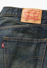 Farkut sinisellä pesulla, joissa on ruskea nahkaleima, johon on merkitty "Levi Strauss & Co." ja punainen logo-lappu takataskussa.