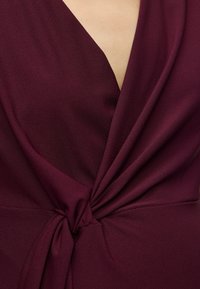 Gros plan d’un tissu bordeaux foncé torsadé et noué à la taille, avec un col en V dévoilant une peau claire.