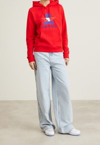 Felpa rossa con un grande logo a stella blu e bianca, abbinata a jeans a gamba larga blu chiaro e scarpe bianche. Stile casual e vestibilità rilassata.