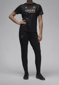 Persona vestida con el uniforme negro del Paris Saint-Germain, con el logotipo de Qatar Airways, de pie con una mano en la cadera contra un fondo liso.