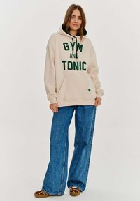 Béžová mikina s kapucí s nápisem "GYM AND TONIC" v zelené, velkým písmem. Obsahuje stahovací kapuci a přední kapsu. V kombinaci s volnými modrými džíny.