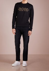 Uomo che indossa un maglione nero Hugo Boss, jeans neri e sneakers bianche, in piedi contro uno sfondo beige uniforme.