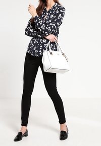 Kobieta ubrana w czarne spodnie i granatową kwiatową bluzkę trzyma białą torebkę, stojąc na tle jednolitego białego tła.