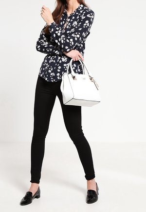 Donna in pantaloni neri e blusa floreale blu navy tiene in mano una borsa bianca, mentre è in piedi di fronte a uno sfondo bianco semplice.