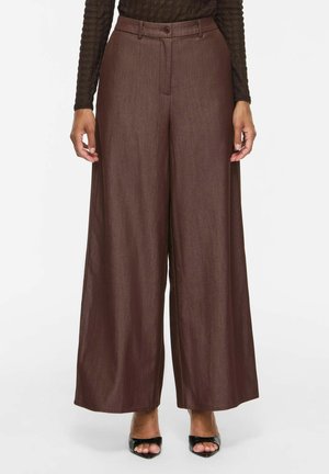 Personne portant un pantalon marron taille haute à jambes larges avec bouton et zip, associé à des talons noirs ouverts et un haut noir à manches longues.