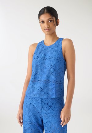 Giovane donna con capelli scuri indossa una canotta di pizzo blu senza maniche e pantaloni abbinati, in piedi davanti a uno sfondo chiaro e uniforme.