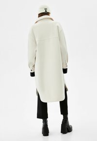 Cappotto beige oversize con colletto, polsini neri a coste e bottoni. Indossato su pantaloni neri e stivali con tacco. Design semplice e strutturato.