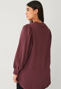 Bordeaux lange mouwen top met opstaande schouders, ronde hals, zijsplitten en een zachte textuur, gezien van achteren.