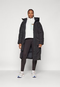Schwarzer, langer Daunenmantel mit Reißverschluss, elastischen Bündchen und Kapuze, getragen über einem weißen Fleece-Oberteil und schwarzen Leggings mit Turnschuhen.