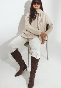 Donna che indossa un maglione beige oversize e una sciarpa, pantaloni bianchi, stivali marroni scuri al ginocchio e occhiali da sole, seduta su una sedia di metallo.