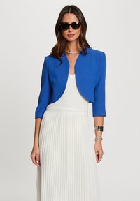 Blauwe cropped jas met driekwartmouwen, gecombineerd met een witte geplooide jurk. Gladde stof, ronde halslijn en minimale hardware.