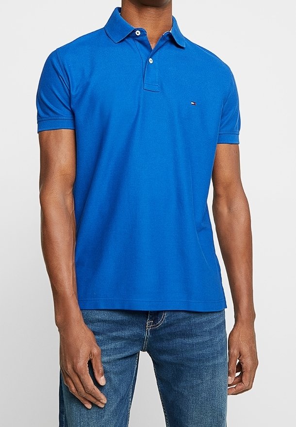 Homme portant un polo à manches courtes bleu vif avec un petit logo sur la poitrine et un jean bleu, debout devant un fond uni.