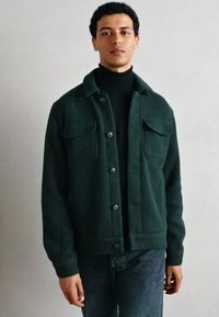 Giacca in velluto a coste verde bosco con quattro tasche, chiusure a bottone e una finitura testurizzata, indossata sopra un dolcevita nero.