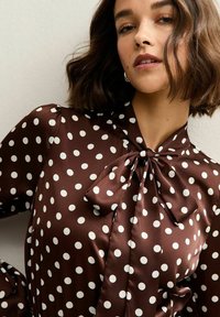 Bruine satijnen blouse met witte stippen, voorzien van een strik bij de kraag en voluminieuze mouwen, met een soepele textuur.