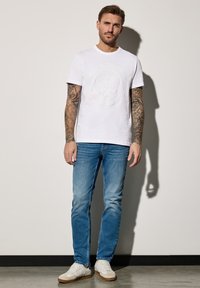 Bílý tričko s krátkým rukávem s reliéfním tenisovým motivem, zkombinované se světle modrými džíny a bílými teniskami. Na rukou jsou viditelné tetování.