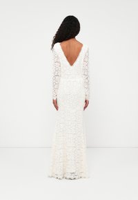 Dolga bela čipkasta obleka z globokim V-izrezom na hrbtu, prilegajočim telesom in razširjenim krilom, ki ima cvetlične vzorce in tekstiurni zaključek.