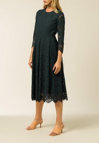 Donkergroen kanten jurk met driekwart mouwen en een golfrand, gecombineerd met lichtbruine open teen hakken. Elegante bloemenpatroon zichtbaar.